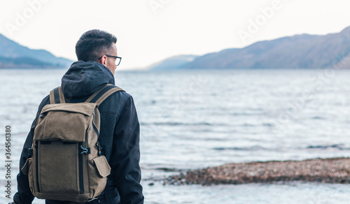 Back view of male hiker in warm jacket with backpack sitting on wooden bench near sea and enjoying marine scenery with rocky coast during travel in Scotland photo