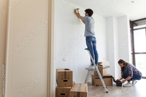 Young man standing on ladder hammering nail and woman picking instruments from bag in apartment with carton boxes photo