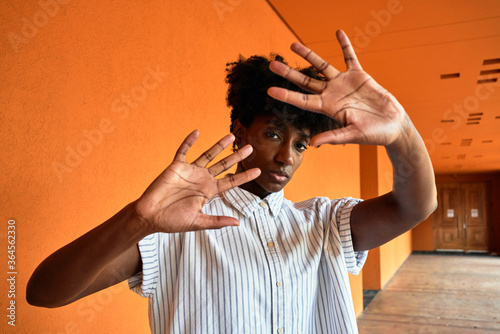 Adult African American female with eyes closed trying to stop negative impact while standing with arms raised against blurred vivid orange interior in corridor of modern building photo