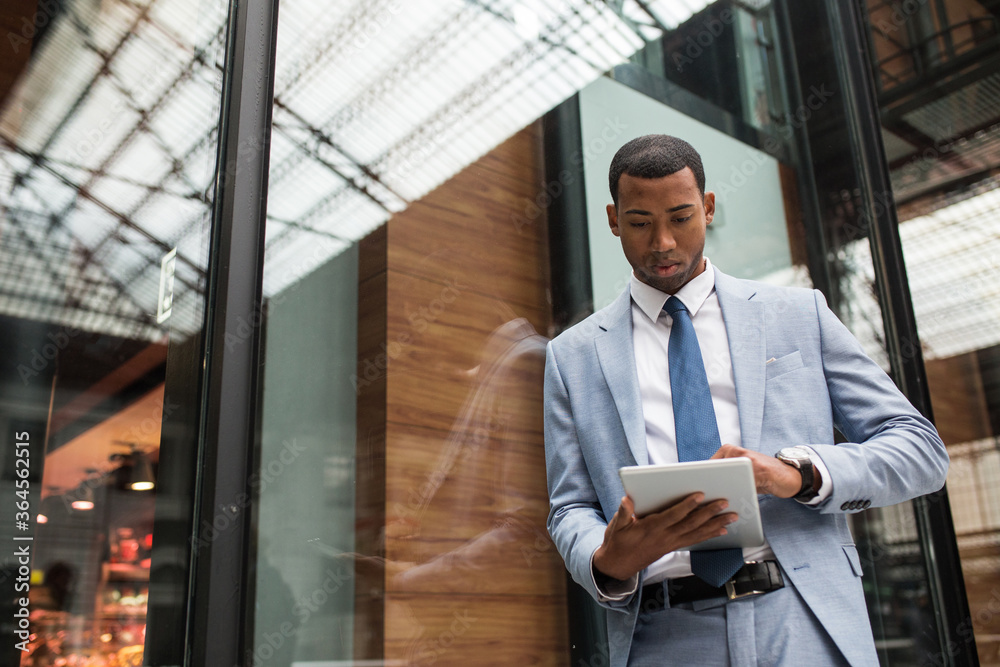 From below view of stylish businessman in elegant suit surfing modern tablet leaning on glass. From below view of stylish businessman in elegant suit surfing modern tablet leaning on glass.
