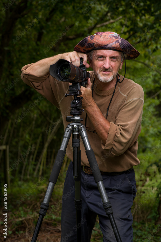 Photographer, mature man, gray beard, floppy hat, brown shirt, blue pants, outside, green wooded area, ready for action from behind DSLR digital camera with zoom telephoto lens on tripod