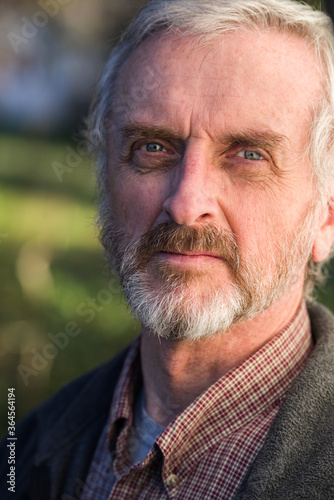 Solemn, serious looking mature man with gray hair and beard outdoors looking towards camera. Checkered shirt and gray sweater outdoors. Portrait.