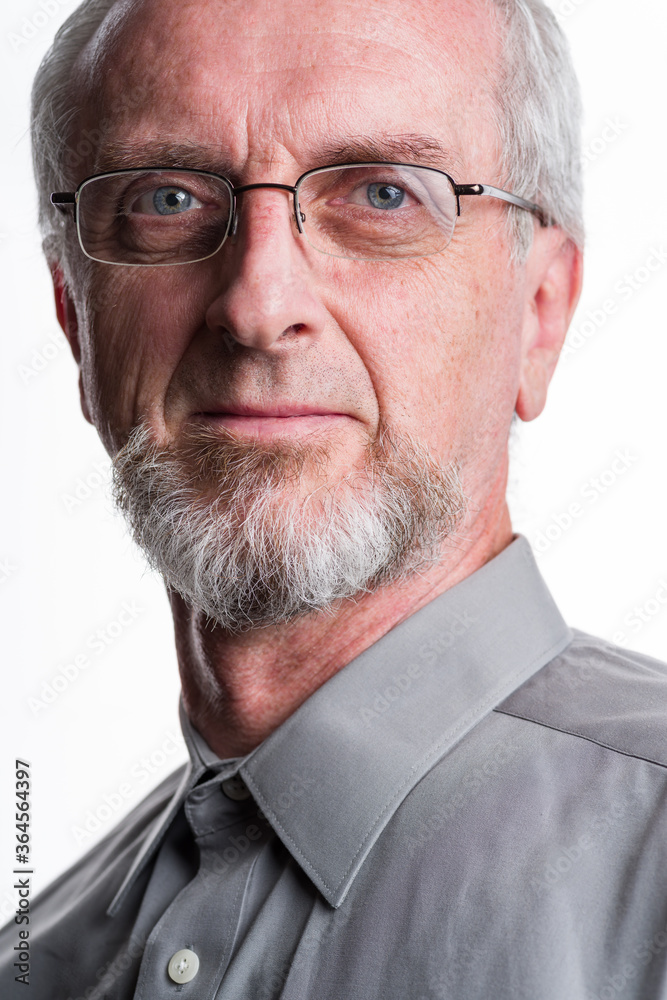 Obraz premium Man in his 50's or 60's with gray grey hair and beard or goatee and button-up gray shirt looking at camera with eye-glasses with confident but tired look in eyes, close-up portrait white background