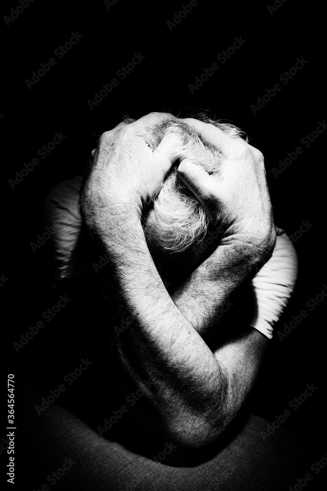 Male depressed, grieving, overwhelmed, alone at table in a dark room covering head and face with hands & arms surrounded by darkness, B&W, black & white high contrast and hard shadows Male depressed, grieving, overwhelmed, alone at table in a dark room covering head and face with hands & arms surrounded by darkness, B&W, black & white high contrast and hard shadows