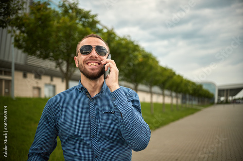 Traveler teen guy calling on the mobile phone. Portrait of confident young businessman dressed in formal clothes standing outside glass building, cell