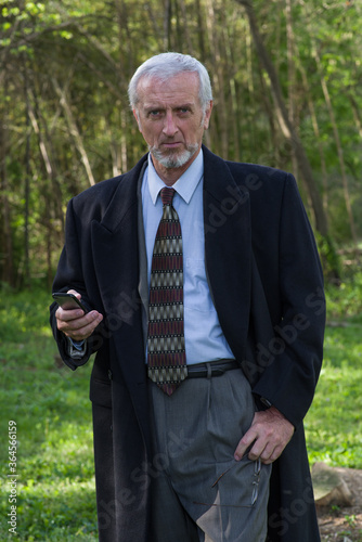 Well dressed older man, suit & tie, long dark wool overcoat, gray hair and beard, three quarter length portrait holding smart phone and with thumb hooked in pocket with green outdoor background