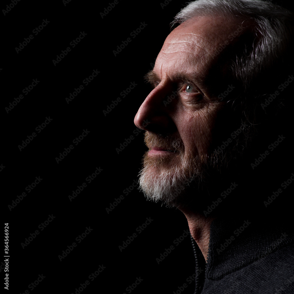 Obraz premium Middle aged man with gray hair and beard, close up profile with hard shadows on black background with eyes open with face and gaze slightly upward in thought, prayer, meditation or contemplation