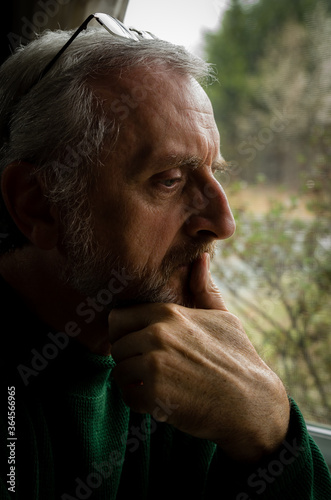 Middle aged man with gray hair and beard, profile gazing out a window with hand to chin in concerned thought or contemplation 