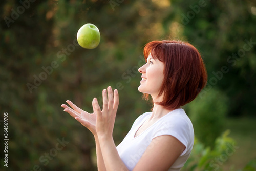 Beautiful woman with red hair catching big green apple falling from above outdoors. Cheerful woman trying to catch apple in garden. Careful planning, success and achieving goals concept © AMR Studio