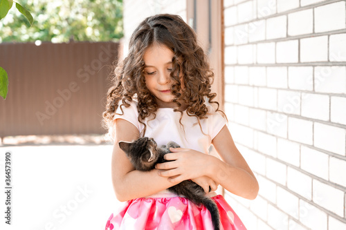 Obraz na płótnie Cute little girl with curls looks at the kitten in her arms with love w salonie