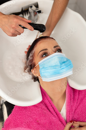 Young woman with surgical mask on face at hairdresser salon. Covid-19 concept. Guy washing beautiful girl's hair at barbershop. High angle view.