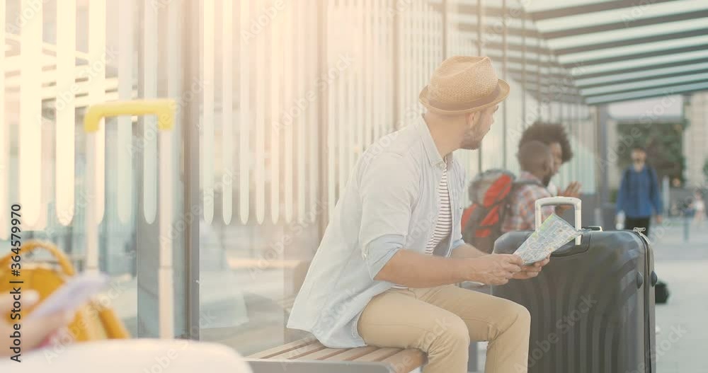 Young Caucasian handsome man traveller in hat sitting on bench on bus stop with city map and waiting for transport. Stylish male tourist looking for route on town plan. Young Caucasian handsome man traveller in hat sitting on bench on bus stop with city map and waiting for transport. Stylish male tourist looking for route on town plan.