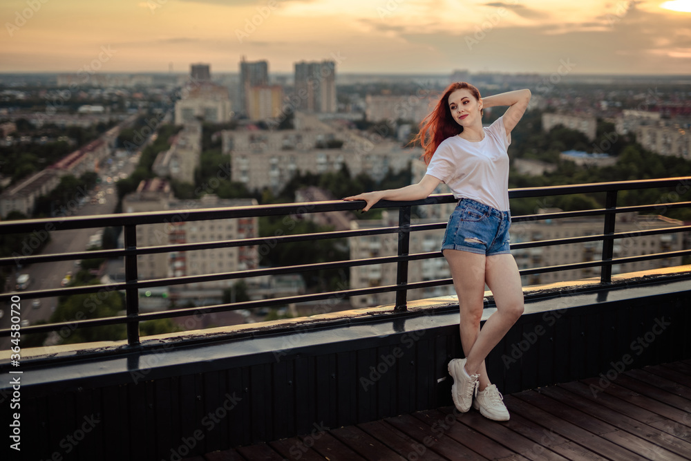 Portrait of a red-haired girl with long hair on the roof of a high house at sunset overlooking the city.