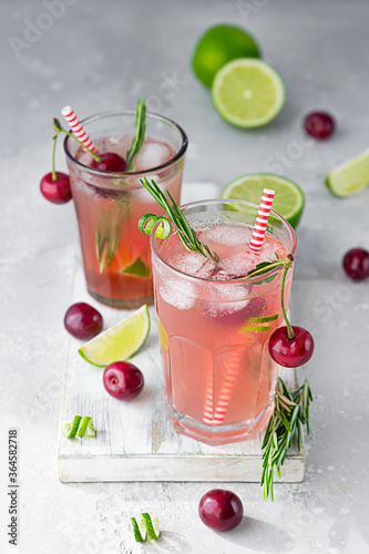 Cold pink non-alcohol cocktail with cherries, lime and rosemary on light wooden cutting board, grey concrete background.