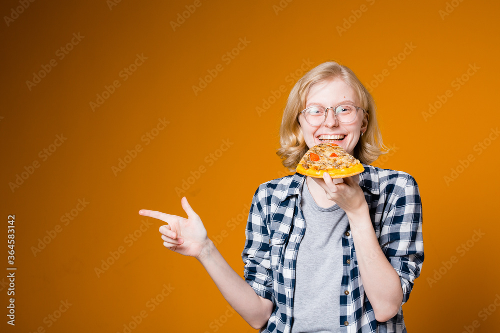 Swedish blonde girl in glasses eating a slice of pizza, smiling and pointing finger to side on an orange background Swedish blonde girl in glasses eating a slice of pizza, smiling and pointing finger to side on an orange background