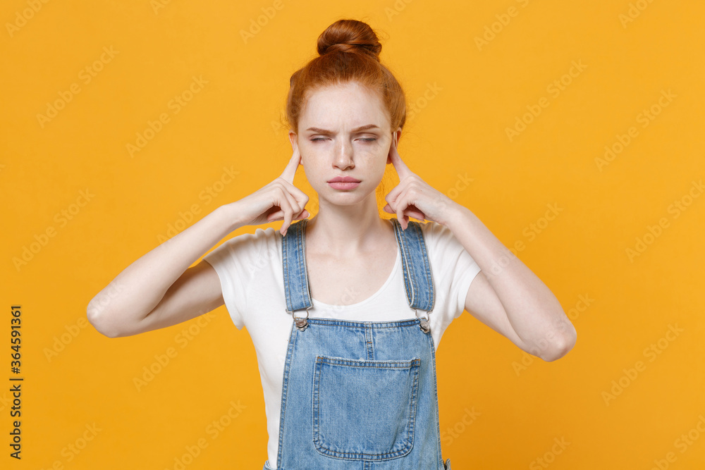 Dissatisfied young readhead girl in casual denim clothes white t-shirt isolated on yellow wall background. People lifestyle concept. Mock up copy space. Covering ears with fingers keeping eyes closed.