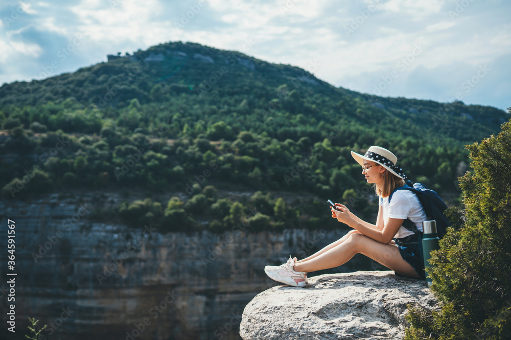 hipster traveler in hat and backpack ralaxing in nature and using internet on smartphone, tourist girl chatting on cellphone on background of panorama horizon mountain landscape copy space