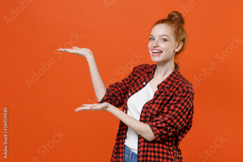 Cheerful young readhead girl in casual red checkered shirt posing isolated on orange background studio portrait. People sincere emotions lifestyle concept. Mock up copy space. Pointing hands aside.