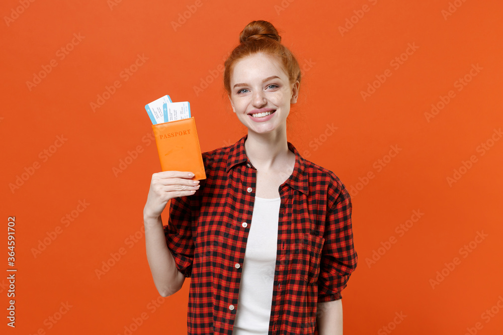Smiling young readhead girl in casual red checkered shirt isolated on orange background. Passenger traveling abroad to travel on weekends getaway. Air flight journey concept. Hold passport tickets. Smiling young readhead girl in casual red checkered shirt isolated on orange background. Passenger traveling abroad to travel on weekends getaway. Air flight journey concept. Hold passport tickets.