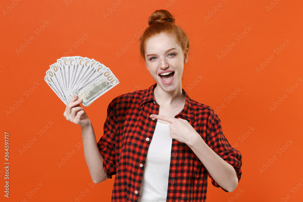 Excited young readhead girl in casual red checkered shirt isolated on orange background. People lifestyle concept. Mock up copy space. Pointing index finger on fan of cash money in dollar banknotes. Excited young readhead girl in casual red checkered shirt isolated on orange background. People lifestyle concept. Mock up copy space. Pointing index finger on fan of cash money in dollar banknotes.