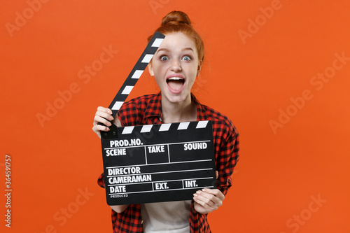 Surprised young readhead girl in casual red checkered shirt posing isolated on orange wall background studio. People lifestyle concept. Mock up copy space. Hold classic black film making clapperboard.