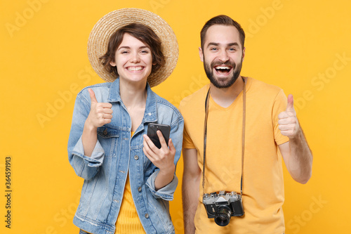 Cheerful young tourists couple friends guy girl with photo camera isolated on yellow background. Passenger traveling abroad on weekends. Air flight journey concept Using mobile phone showing thumb up.