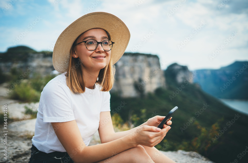 smiling tourist woman in hipster glasses and hat using smartphone and relaxing while walking on tops of mountains, blonde girl traveler with mobilephone hiking summer outdoors enjoys leisure on nature