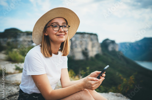 Obraz na płótnie smiling tourist woman in hipster glasses and hat using smartphone and relaxing while walking on tops of mountains, blonde girl traveler with mobilephone hiking summer outdoors enjoys leisure on nature w salonie