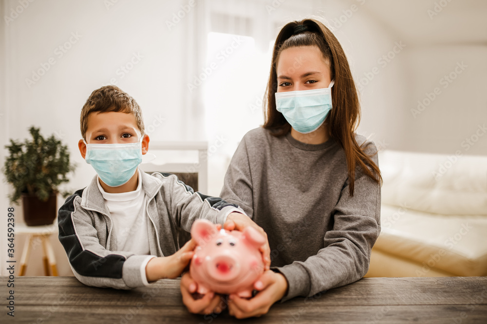 Boy and girl, wear protective masks, holding piggybank at home.