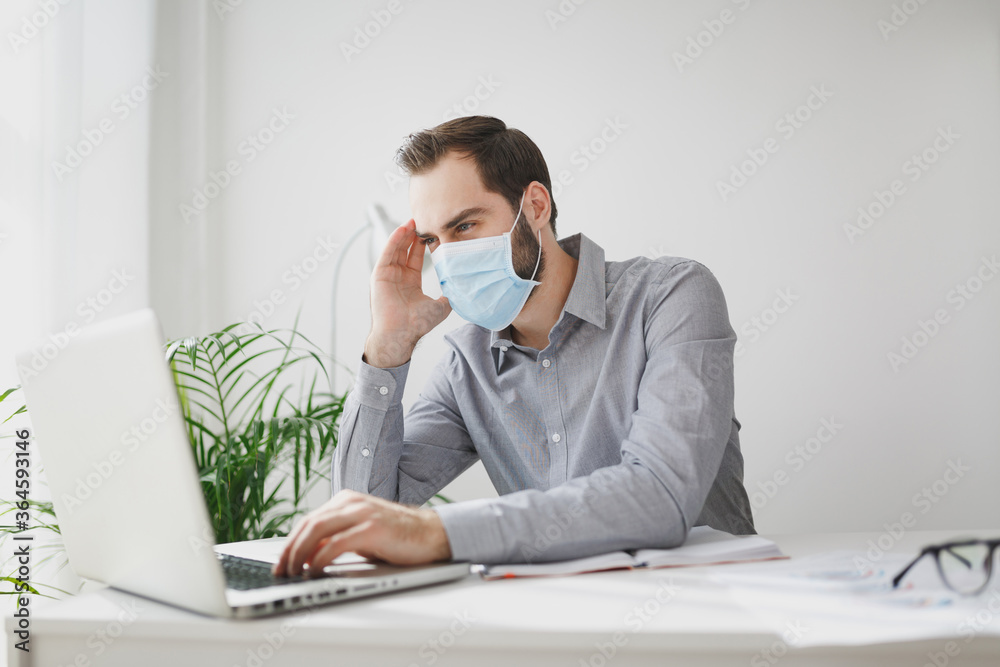 Young bearded business man in gray shirt sterile face mask sitting at desk in light office on white wall background. Achievement business career concept. Work on laptop pc computer put hand on head. Young bearded business man in gray shirt sterile face mask sitting at desk in light office on white wall background. Achievement business career concept. Work on laptop pc computer put hand on head.