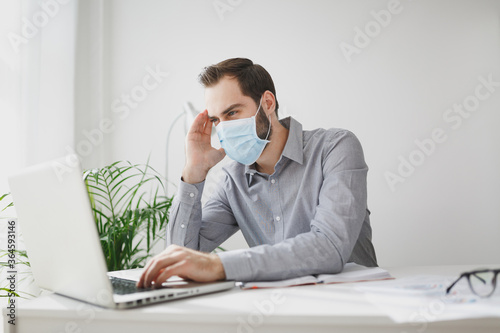 Young bearded business man in gray shirt sterile face mask sitting at desk in light office on white wall background. Achievement business career concept. Work on laptop pc computer put hand on head.