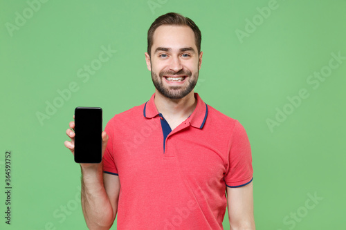 Smiling young bearded man guy in casual red pink t-shirt posing isolated on green background studio portrait. People lifestyle concept. Mock up copy space. Hold mobile phone with blank empty screen.