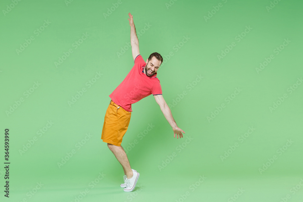 full length Cheerful young man guy in casual red pink t-shirt posing isolated on green background studio portrait. People lifestyle concept. Mock up copy space. Stand on toes, spreading hands, dancing full length Cheerful young man guy in casual red pink t-shirt posing isolated on green background studio portrait. People lifestyle concept. Mock up copy space. Stand on toes, spreading hands, dancing