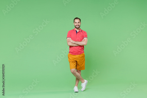 full length shot Smiling young bearded man guy in casual red pink t-shirt posing isolated on green wall background studio portrait. People lifestyle concept. Mock up copy space. Holding hands crossed. © ViDi Studio