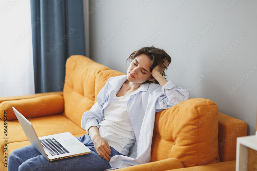 Tired young girl in casual clothes sit on couch spending time in living room at home. Rest relax good mood leisure lifestyle concept. Work on laptop pc computer, put hand on head, keeping eyes closed. Tired young girl in casual clothes sit on couch spending time in living room at home. Rest relax good mood leisure lifestyle concept. Work on laptop pc computer, put hand on head, keeping eyes closed.