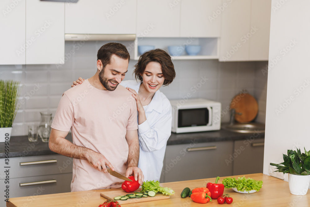 Smiling young couple two friends guy girl in casual clothes preparing vegetable salad cooking food in light kitchen at home. Dieting family healthy lifestyle concept. Mock up copy space. Hugging.