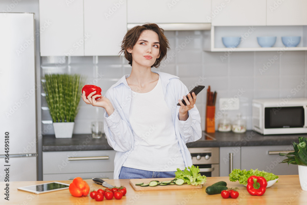 Pensive housewife woman in casual clothes using mobile phone looking for recipe preparing vegetable salad cooking food in light kitchen at home. Dieting healthy lifestyle concept. Hold bell pepper.