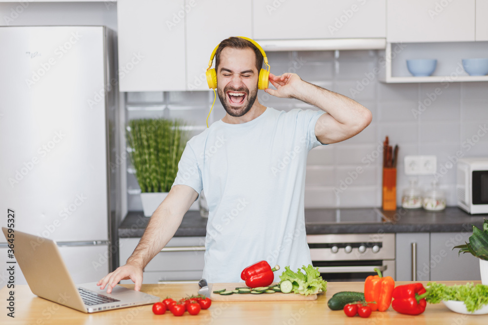 Cheerful young bearded man in casual t-shirt with headphones using laptop computer looking at recipe preparing vegetable salad cooking food in light kitchen at home. Dieting healthy lifestyle concept. Cheerful young bearded man in casual t-shirt with headphones using laptop computer looking at recipe preparing vegetable salad cooking food in light kitchen at home. Dieting healthy lifestyle concept.