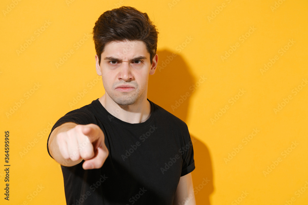 Dissatisfied young man guy in casual black t-shirt posing isolated on yellow background studio portrait. People sincere emotions lifestyle concept. Mock up copy space. Pointing index finger on camera.