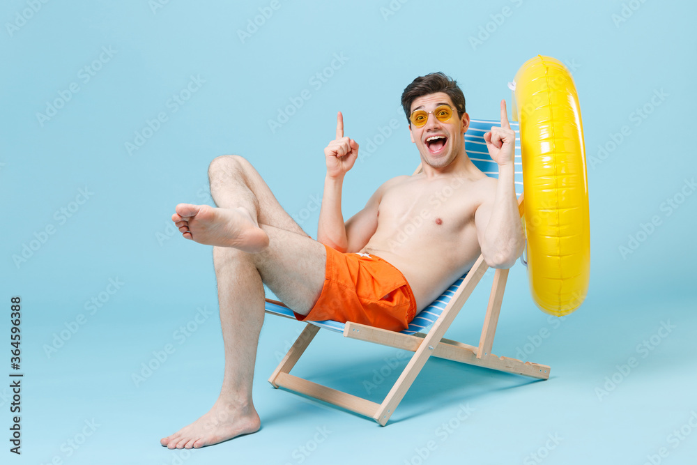 Surprised young man in orange shorts glasses sit on deck chair isolated on blue wall background studio. People summer vacation rest lifestyle concept. Mock up copy space. Pointing index fingers up. Surprised young man in orange shorts glasses sit on deck chair isolated on blue wall background studio. People summer vacation rest lifestyle concept. Mock up copy space. Pointing index fingers up.