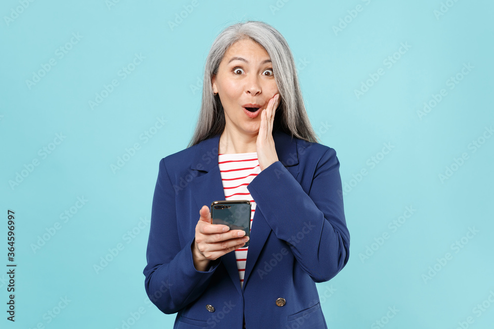 Shocked gray-haired business woman in blue suit posing isolated on pastel blue background studio. Achievement career wealth business concept. Mock up copy space. Using mobile phone, put hand on cheek. Shocked gray-haired business woman in blue suit posing isolated on pastel blue background studio. Achievement career wealth business concept. Mock up copy space. Using mobile phone, put hand on cheek.