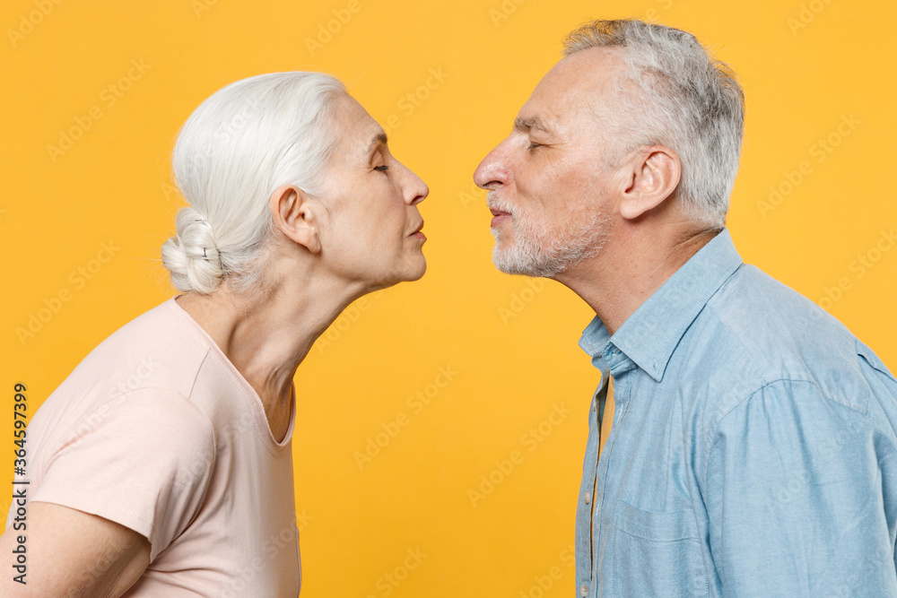 Side view of pretty elderly gray-haired couple woman man in casual clothes posing isolated on yellow wall background studio. People lifestyle concept. Mock up copy space. Kissing, keeping eyes closed. Side view of pretty elderly gray-haired couple woman man in casual clothes posing isolated on yellow wall background studio. People lifestyle concept. Mock up copy space. Kissing, keeping eyes closed.