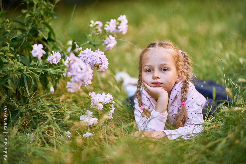The little girl lies on the green grass near the bush with pink flowers and makes different facial expressions. girl in pink blouse and blond hair The little girl lies on the green grass near the bush with pink flowers and makes different facial expressions. girl in pink blouse and blond hair