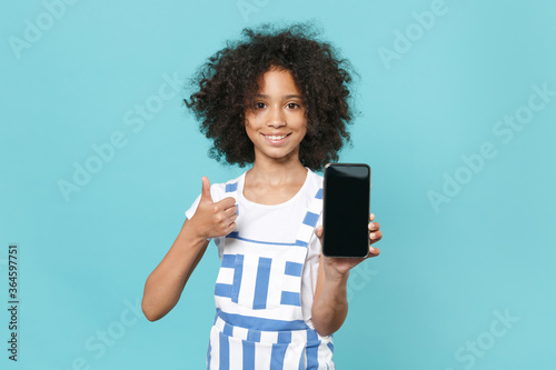 Smiling little african american kid girl 12-13 years old in striped clothes isolated on blue wall background. Childhood lifestyle concept. Hold mobile phone with blank empty screen, showing thumb up.