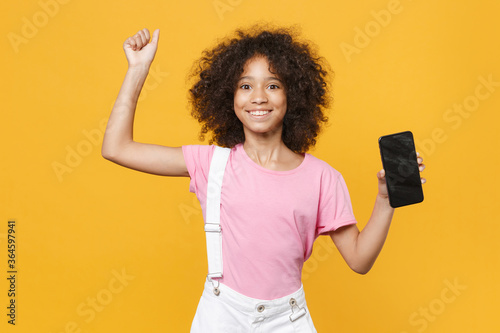 Joyful african american kid girl 12-13 years old in pink t-shirt isolated on yellow background. Childhood concept. Mock up copy space. Hold mobile phone with blank empty screen, doing winner gesture.