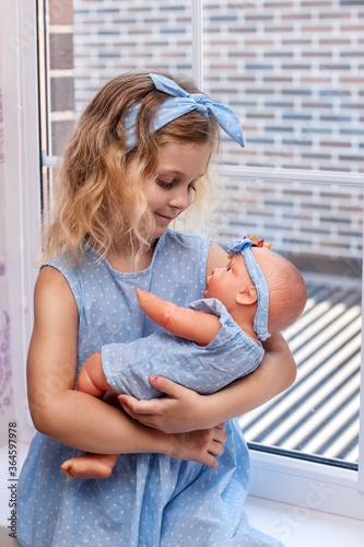 a charming blonde six year old girl in a blue dress holds a doll in her hands sitting on the windowsill of her bedroom