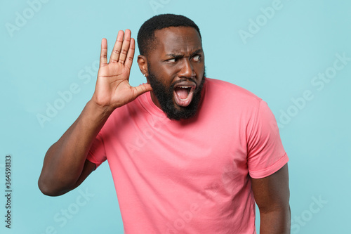 Shocked young african american man guy in casual pink t-shirt posing isolated on blue wall background studio portrait. People lifestyle concept. Mock up copy space. Try to hear you listening intently.