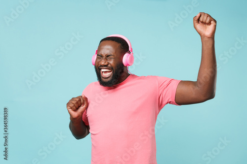 Funny young african american man guy in casual pink t-shirt isolated on blue background studio. People lifestyle concept. Mock up copy space. Listen music with headphones keeping eyes closed, dancing.