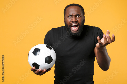 Angry african american man football fan in black t-shirt isolated on yellow background. Sport family leisure lifestyle concept. Cheer up support favorite team with soccer ball, swearing, screaming.