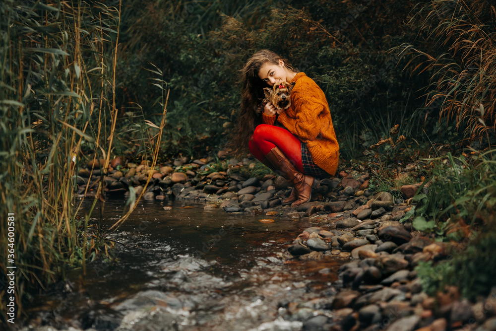 a girl with long hair and an orange sweater and red tights holds a dog in her arms. In the open air by the stream. a girl with long hair and an orange sweater and red tights holds a dog in her arms. In the open air by the stream.